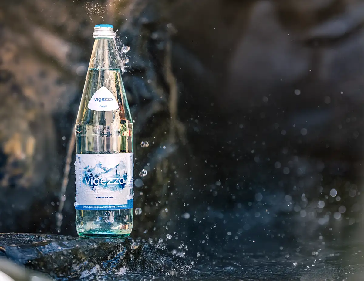 Eine Flasche aus klarem Glas mit Vigezzo Mineralwasser steht auf einem Stein im Wasser und wird angespritzt.